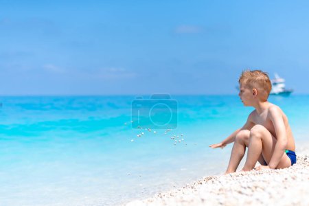 Téléchargez les photos : Un garçon joue avec des cailloux sur la belle plage de galets près de l'eau turquoise - en image libre de droit