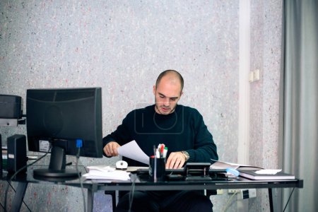 Homme d'affaires multi-tâches à son bureau, assis dans son bureau et travaillant avec des papiers et en ligne aussi.