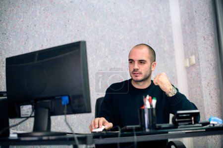 Jeune homme d'affaires assis à son bureau, regardant le moniteur d'ordinateur et le succès gestuel.