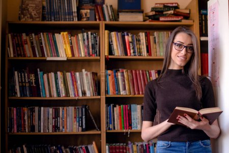 Junge Frau in Bibliothek, Buch in der Hand und lächelnd.