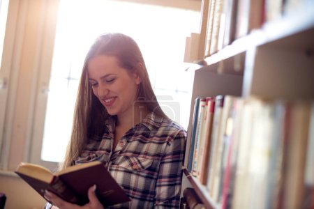 Smiling young girl in library, standing next to bookshelf and reading a book.