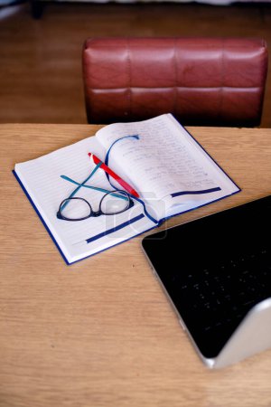 Photo for Cropped image of office tools at wooden desk: laptop, notebook and eyeglasses, office chair also. - Royalty Free Image