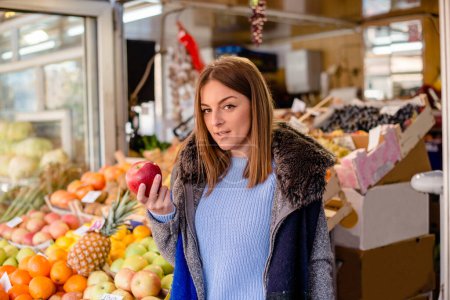 Photo for Beautiful woman standing at greengrocer's shop, holding red apple. - Royalty Free Image