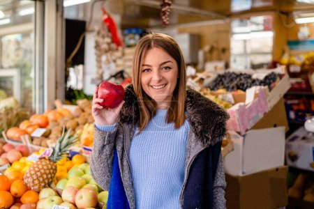 Photo for Portrait of smiling woman holding red apple, standing at market, orange and pineapple at market stall. - Royalty Free Image