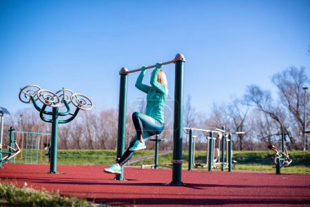 Woman exercising on sports utensils in park, doing chin ups.
