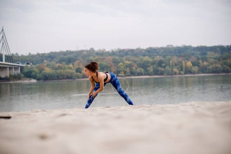 Atleta mujer cansada terminando su entrenamiento, estirándose en la arena, mirando hacia otro lado.
