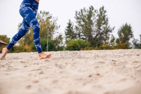 Woman running on the beach, wearing blue jogging pants and barefoot - low section.