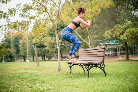 Photo for Woman jumping in park, doing squats on park bench. - Royalty Free Image