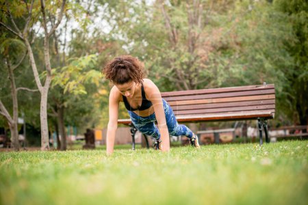 Young woman doing push-ups on grass in public park.