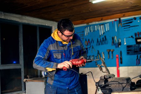 Man using power drill in workshop, wearing protective wear and eyeglasses.