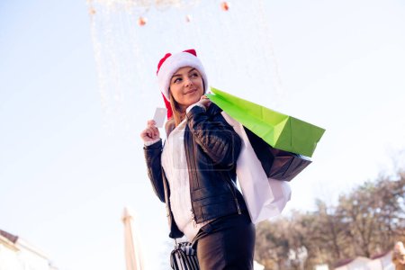 Téléchargez les photos : Jeune femme avec sac à provisions, tenant la carte de crédit dans sa main, portant le chapeau de Père Noël. - en image libre de droit