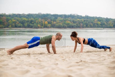 Man and woman doing push-ups on sand, river and green plants in background.