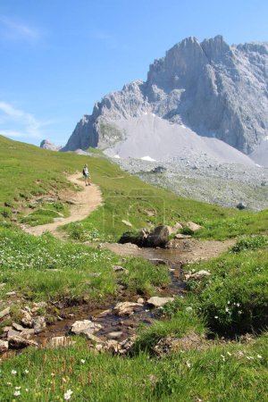 Wanderweg mit Flüsschen und Wanderinnen im Rücken, Weg zum SAC Carschina Schweiz Wandersaison Sommer mit wenig Schnee. Hochwertiges Foto
