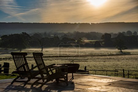 Foto de Reclinando sillas, sentarse y relajarse en una hermosa vista del campo de un glorioso amanecer sobre el paisaje rural herboso en Devon Reino Unido. Ganado pastando en la distancia con una niebla colgando sobre los campos. - Imagen libre de derechos