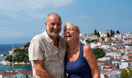 Téléchargez les photos : Couple heureux riant pendant les vacances. Capture détendue, amusante et ensoleillée d'un homme et d'une femme d'âge moyen partageant une blague avec une toile de fond de maisons blanchies en terre cuite. - en image libre de droit