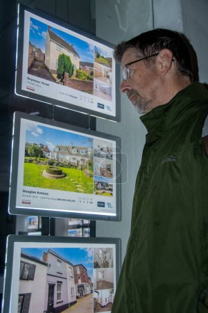 Foto de Exmouth UK. 10-13-23. Inmobiliaria ventana publicidad propiedades en venta o alquiler. Un hombre en busca de un nuevo hogar navega por el High St por la noche. - Imagen libre de derechos