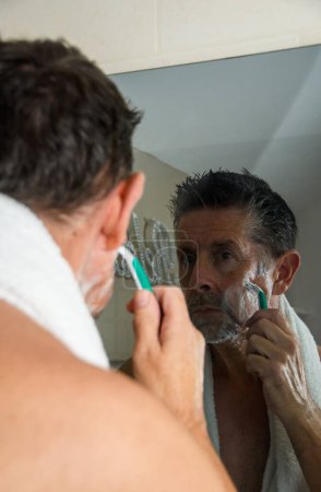 Téléchargez les photos : Un homme se rasant dans le miroir, son reflet est capturé dans une salle de bain sombre. - en image libre de droit