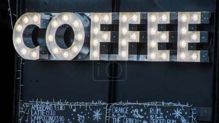 Téléchargez les photos : Café dans un marché de Noël. Fond noir avec de grandes lumières blanches illuminées annonçant des thés chauds et des boissons au café à vendre auprès d'un vendeur indépendant sur une place du marché. - en image libre de droit