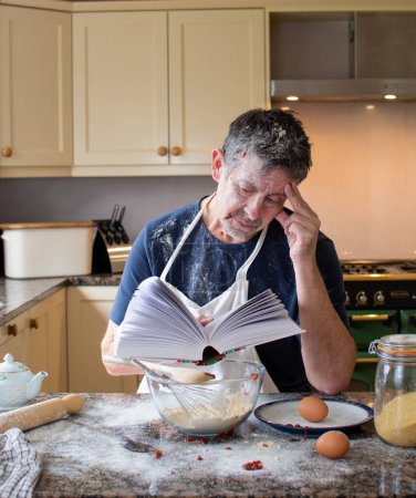 Photo for Man looks at a cookery book in the kitchen. Wearing an apron, surrounded by ingredients and mess. Learning how to bake is a new challenge for a mature man. Real life skills concept. Cookery school. - Royalty Free Image