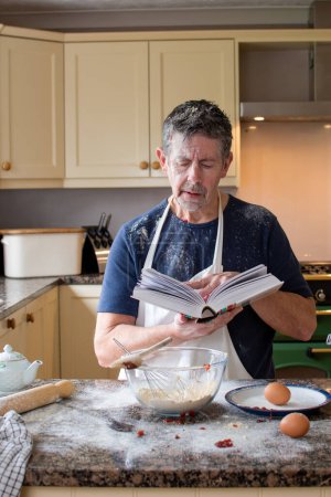 Photo for Man looks at a cookery book in the kitchen. Wearing an apron, surrounded by ingredients and mess. Learning how to bake is a new challenge for a mature man. Real life skills concept. Cookery school. - Royalty Free Image