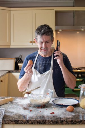 Photo for Man calls for help to bake a cake, holding an egg in one hand and a mobile phone in the other, he's no MasterChef. Dad is attempting a Bake off, he's confused and making a mess. Funny capture. - Royalty Free Image