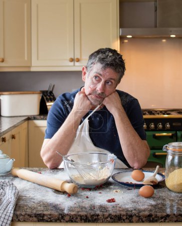 Photo for Failure at baking a cake in cookery lessons. Senior man Looking fed up and miserable at his attempt to create a meal. Kitchen disaster, with flour and jam all over the kitchen. - Royalty Free Image