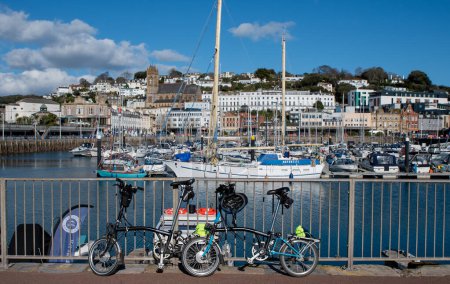 Téléchargez les photos : Torquay, Devon, Royaume-Uni. 02-12-24. Port de Torquay, deux vélos pliants Brompton sur fond de ville lumineuse et de marina. Casques, vestes l'image complète de vacances de vélo de tourisme concept. - en image libre de droit
