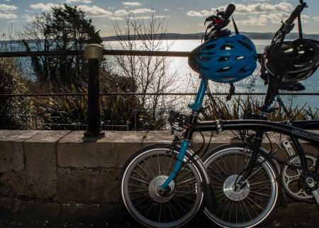 Téléchargez les photos : Deux vélos pliants Brompton sur fond de port et de bord de mer lumineux. Casques, vestes et sacs à dos complètent l'image du concept de vacances à vélo de randonnée. - en image libre de droit