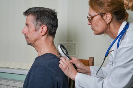 Doctor examining a patient at an appointment. A female GP wearing a white coat checks her patient's skin with a magnifying glass for signs of cancer, whilst discussing treatment at the hospital clinic