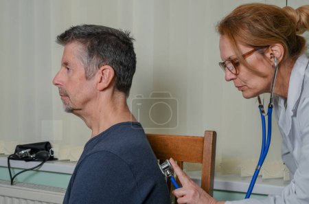 Doctor examining a patient at an appointment. A female GP wearing a white coat checks her patient's lungs for crackles with a stethoscope whilst discussing the treatment at the hospital or clinic.