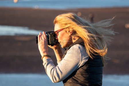 Photo for Photographer image. A woman with a DSLR takes an outdoor picture on a windy day. Her hair blows in the wind as she focuses the camera. Warm light at sunset casts across her. - Royalty Free Image