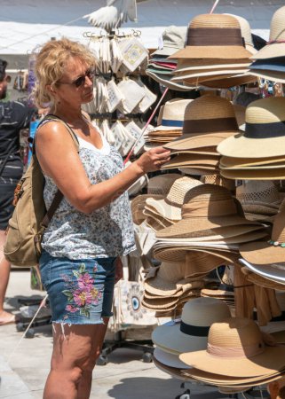 Photo for Skiathos, Greece. 07-06-23. A lady browses hats in the hot Greek sun. Skiathos tourism and commerce picture. Selective focus on the ladies face. - Royalty Free Image