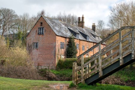 Photo for Scenic red brick mill house. Beautiful old building situated on a riverside with wooden steps in the foreground. Outstanding rural country image. Country lifestyle and rural home picture. Editorial. - Royalty Free Image