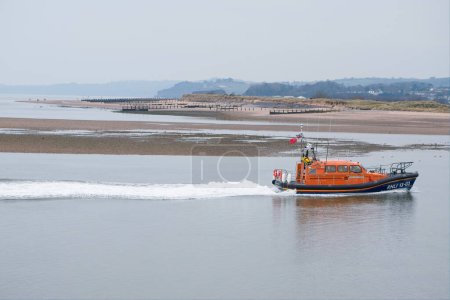 Photo for Exmouth, Devon, uk. March 5th 2023. Exmouth lifeboat on a flat calm sea with Dawlish warren behind. A long wake ripples the flat water behind the boat. Focus on the lifeboat. Copy space. - Royalty Free Image