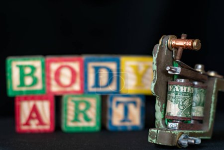 Photo for Churston, devon, uk. December 17th 2022. Tattoo gun with selective focus on the word American. Wooden letter blocks say the words body art in soft focus. Body art image. Tattoo artist. - Royalty Free Image