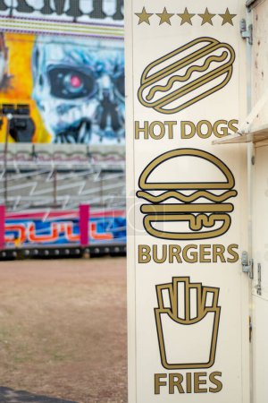Photo for Paignton, Devon, uk. August 23rd 2022. Paignton fairground fast food sign. Burgers, hot dogs, fries sign in selective focus. Fairground ride in soft focus behind. Beige metal board with food graphics - Royalty Free Image
