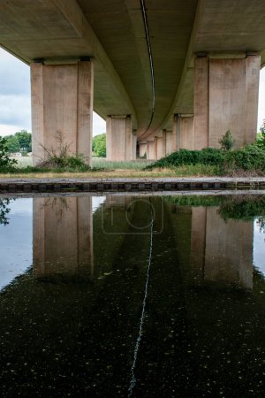 Photo for A bridge from underneath reflected in water. A dividing line between the two carriageways creates a stark white line meandering as a reflection across the dark black water. Strongly contrasting image. - Royalty Free Image