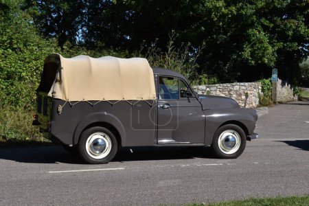 Photo for Powderham, Devon, uk. July 9th 2022. A classic Morris Minor pickup with a tan canvas roof drives down a country lane on a sunny afternoon. White steel wheels show off this rare historic vehicle. - Royalty Free Image