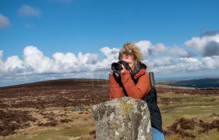 Photo for Dartmoor national park. A woman with a DSLR camera leans against a granite post on Dartmoor as she takes a photograph. She wears warm winter clothing to protect her from the cold moorland weather. - Royalty Free Image