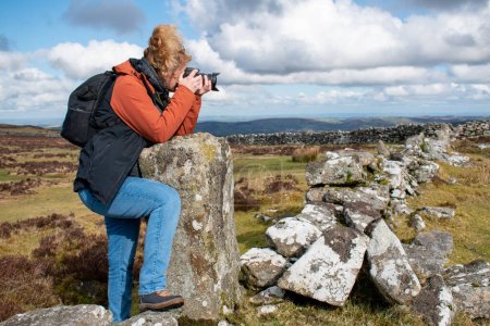 Photo for Dartmoor national park image. A side profile picture of a woman with a DSLR camera taking a photograph on a cold winters day on Dartmoor National Park. Granite walls and moorland landscape fill the scene - Royalty Free Image