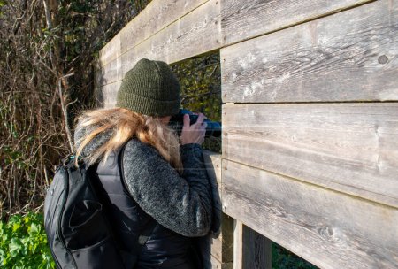 Photo for Exeter, devon, england, 08 January 2022. Woman photographer with DSLR camera takes a picture at a bird hide on a winters day. Long lens to capture birds . Woolly hat and thick warm fleece. - Royalty Free Image