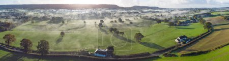 Téléchargez les photos : Vue aérienne panoramique de la campagne près de Honiton et Ottery St Mary. Lever de soleil avec éclat de lentille et brouillard roulant traversent les champs verts luxuriants ci-dessous. Paysage spectaculaire. - en image libre de droit