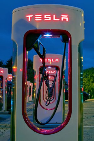 Foto de Exeter, Devon, Reino Unido. 24-10-23. Tesla EV estaciones de carga en una línea por la noche. Rojo Tesla signos que brillan con el poder. Enfoque selectivo en la primera estación de carga. - Imagen libre de derechos