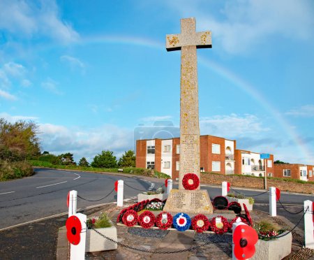 Photo for Budleigh Salterton, uk. 11-28-23. Poppies at a war memorial with a vivid rainbow on a perfect blue sky behind. Names of the fallen sit at the base of a stone cross. Memorial for veterans. - Royalty Free Image