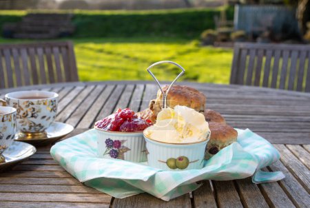 Bollos con tazas de té en un jardín inglés. Completo con mermelada de fresa fresca y crema coagulada Devonshire. Luz solar moteada con pastos verdes más allá. Enfoque selectivo en la jam pot.