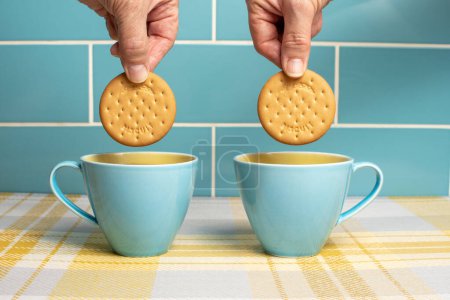 Cups of tea in attractive blue and yellow cups with hands dunking rich tea biscuits. Cups isolated against a blue tiled background and sitting on yellow check. Mugs of hot drink with a sweet treat. 