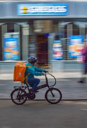 Photo for Exeter, Devon, uk. 03-21-24. Food delivery image. Deliveroo and Just eat. Fast food delivered by cyclist taken with motion blur. Greggs fast food shop blurred behind. - Royalty Free Image