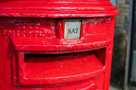 Palabra del sábado mostrada como SAT en un buzón rojo británico. Imagen en formato retrato visualmente llamativa y colorida que representa el día de la semana. Rojo brillante. Buzón e imagen postal. 