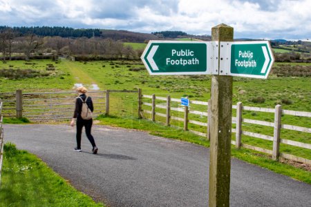 Country walks and public footpath image. A signpost marked as public footpath in sharp focus with a softly focused lady walking the path behind. Rural countryside and exercise. 