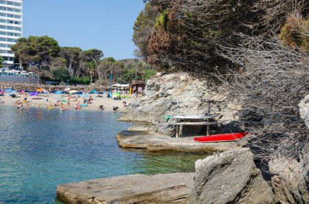 Photo for Beach with tourists and red boat in summer in spain. On the left are rocks, trees and dry branches. - Royalty Free Image